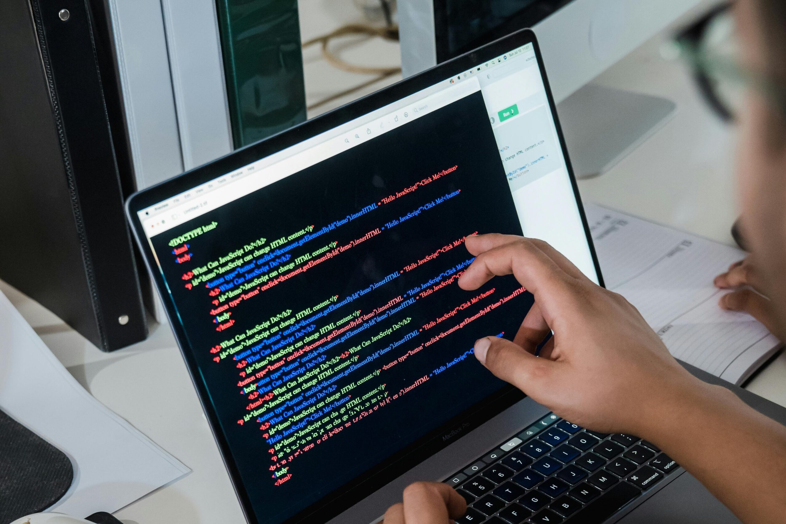 Close-up of a programmer pointing at a colorful code script on a laptop in an office setting.